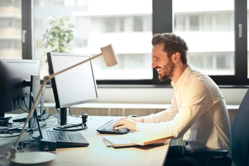 man-working-at-desk A man smiling while working at an office desk with a computer and natural daylight streaming in through large windows.